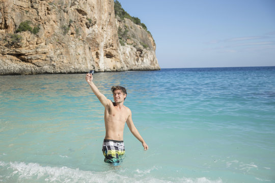 Young man in ocean arm raised using camera smiling, Golfo di Orosei, Sardinia, Italy