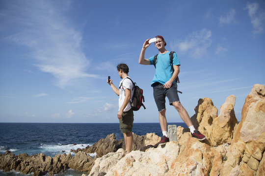 Young men standing on rocks using smartphone to take photograph, Costa Paradiso, Sardinia, Italy
