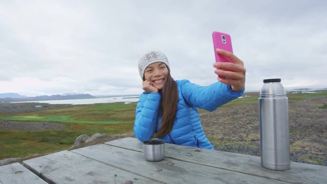 Woman outdoors using smart phone taking selfie photo on app on smartphone while sitting outside wearing warm down jacket. Mixed race Asian Chinese Caucasian woman in active lifestyle, Iceland.