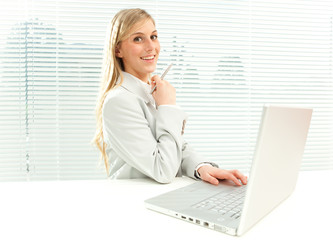 smiling blond businesswoman with laptop near venetian blind window