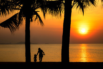 Beautiful sunset on beach with caring happy mother and daughter walking under coconut tree in silhouette twilight golden hour/Sunset on beach, happy mother and daughter
