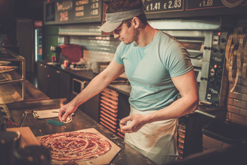 Handsome pizzaiolo making pizza at kitchen in pizzeria. - obrazy, fototapety, plakaty