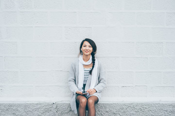 Young beautiful asiatic woman sitting leaning against a white wall, listening music with earphones...