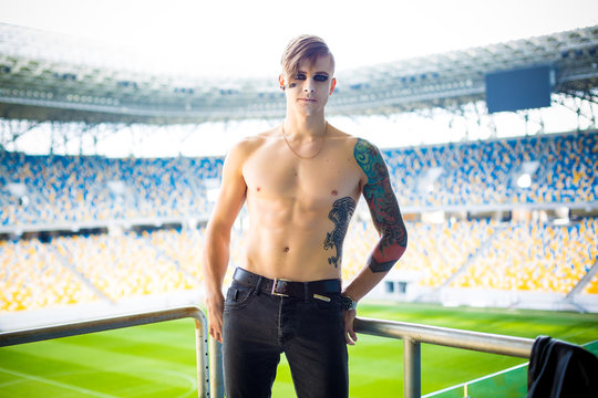Portrait of tattooed ill boy with short hair and facial stubble wearing a jacket, black jeans and posing for stli football stadium. Hipster style.