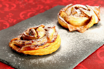 Two little roses of puff pastry with apples, cinnamon and powdered sugar on a slate dish. Red background.