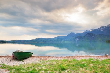 Abandoned fishing paddle boat on bank of Alps lake. Morning lake glowing by sunlight.