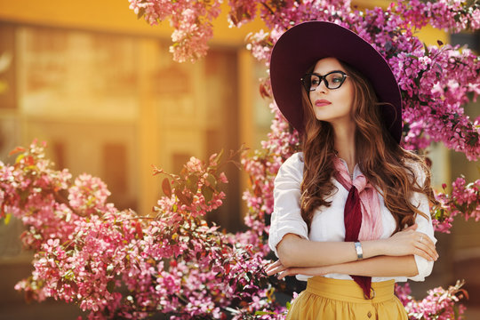 Outdoor portrait of young beautiful fashionable lady posing near flowering tree. Model wearing stylish accessories & clothes. Girl looking aside. Female beauty & fashion concept. City lifestyle. Toned