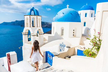 Santorini travel tourist woman on vacation in Oia walking on stairs. Person in white dress visiting the famous white village with the mediterranean sea and blue domes. Europe summer destination. - obrazy, fototapety, plakaty
