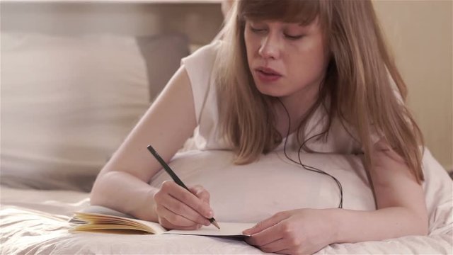 Attractive young woman lying on the bed with the phone listening to music, studying and taking notes in a book 