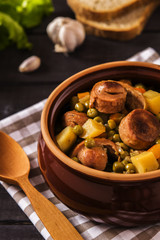 Vegetable stew with pork sausages on dark beer in ceramic bowl on a dark wooden background with garlic, herbs and bread 