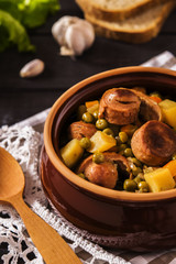 Vegetable stew with pork sausages on dark beer in ceramic bowl on a dark wooden background with garlic, herbs and bread 