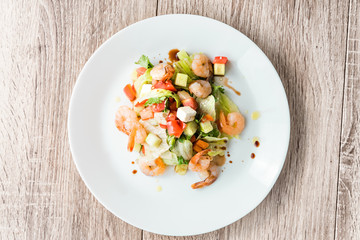 Vegetable salad with shrimps and feta cheese on a white plate on a wooden table, top view