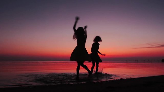 Silhouettes of two happy girls whirling and dancing at the beach on sunset