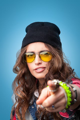 Closeup studio portrait of beautiful trendy hipster girl with long curly hair pointing finger at you wearing  red checkered shirt, denim vest,yellow glasses and black beanie hat.
