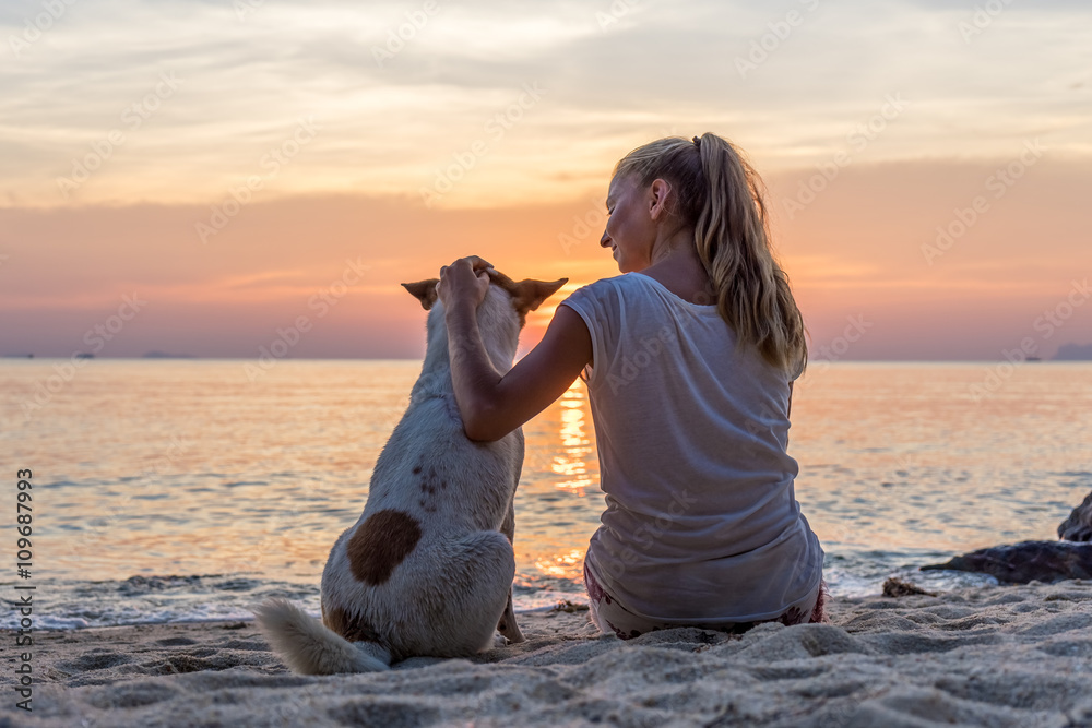 Wall mural Young woman with dog sitting on the beach and watching the sunset
 - Wall murals