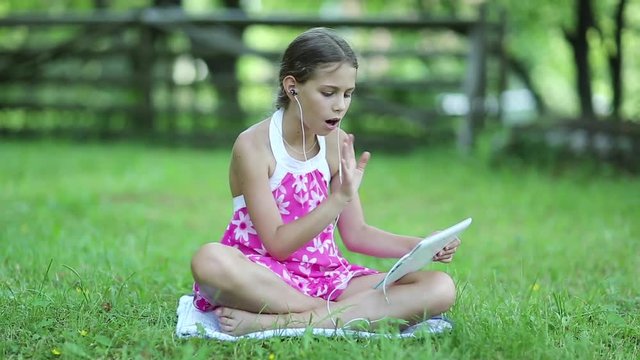 Beautiful girl in pink dress with tablet computer sits on green grass in garden. Little girl with tablet pc sits on the grass in garden. Girl communicates through Skype. Female with tablet computer