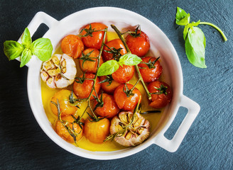 Small cherry tomatoes and garlic baked with salt and olive oil. White baking dish on black stone background. Top view.