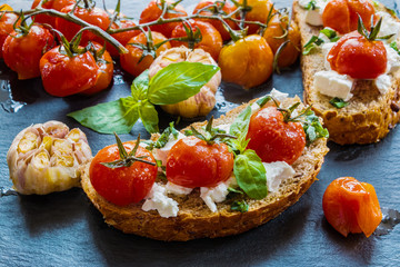 Toasts with feta cheese and small cherry tomatoes baked with garlic, salt and olive oil. Black stone background.