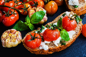 Toasts with feta cheese and small cherry tomatoes baked with garlic, salt and olive oil. Black stone background.