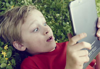 Portrait of Child blond young boy playing with a digital tablet computer outdoors  lying on grass, listening music or watching movie, facial expression