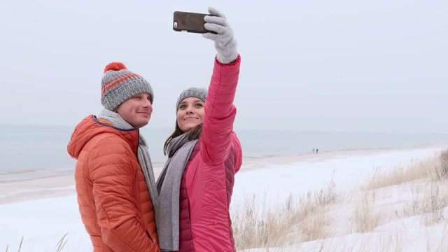 Couple taking a selfie at the beach in winter, girl is flirting with the camera and kissing the guy who is so cold. Medium shot.
