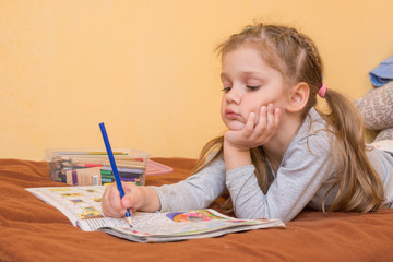 Little girl studying a magazine with a pencil in his hand lying on his stomach and his head in his second hand