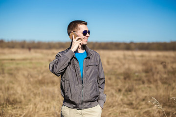 Young man with sunglasses and windbreaker talking on the mobile phone. There is a  field as background