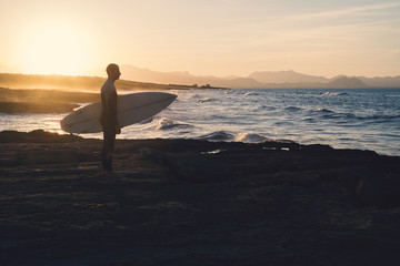 Young man surfer holding the surfboard while contemplating the waves at sunset.