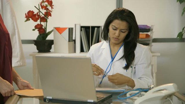 A nurse brings more paperwork to a fatigued female physician working in her office.