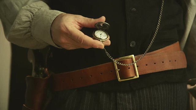 A gunslinger from the American wild west era checks the time on his pocket watch and then his pistol.