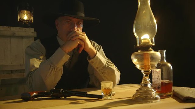 A mature pensive cowboy from the American wild west era sitting with booze and pistol contemplates trouble.