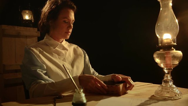 A pensive woman from the era of the early American west slowly opens her Bible and begins to read by the light of an old oil lamp.