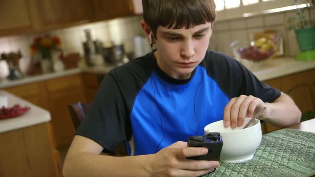 A teenage boy eating cereal listens to music as he reads text messages on his smart phone.