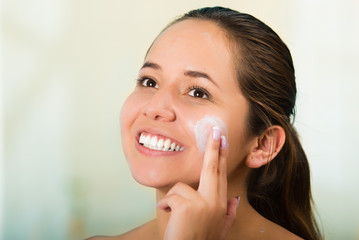 Pretty young healthy hispanic woman headshot with naked shoulders, smiling happily and applying cream to face using hands
