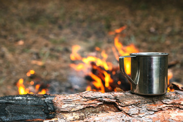 Iron cup with hot tea stands on a log near the fire. Journey into the wild concept.