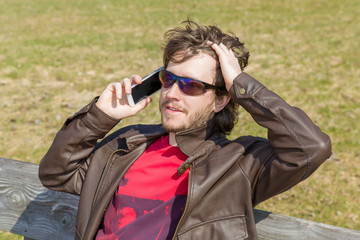 Young man wearing brown leather jacket and sunglasses, holding mobile phone, making a call and speaking on the phone and resting in the park in spring day.