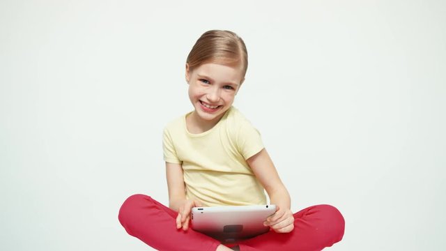 Girl 7-8 years old using tablet pc and sitting on the floor on white background and smiling at camera