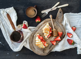 Breakfast set. Freshly baked croissants with strawberries, mascarpone, honey and coffee on rustic wooden board over dark grunge backdrop