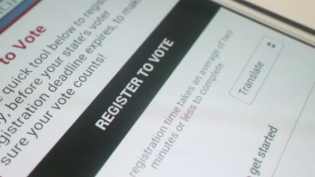 Close up overhead dolly to tablet-phablet, settling on black banner that reads 'Register to Vote'. User finger then presses banner, with camera dolly back out to 1st position. 
