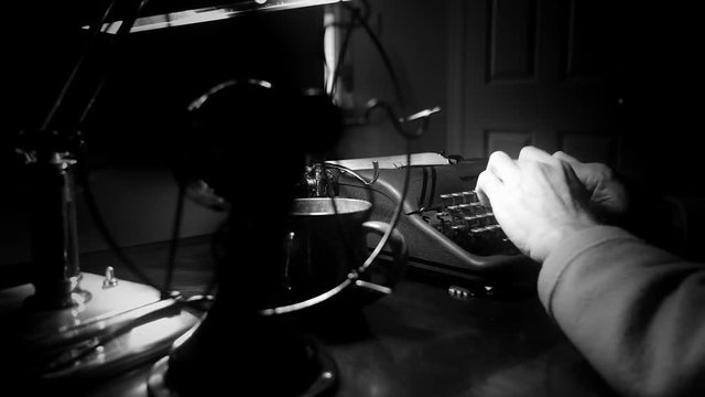 Through the turning blades of a vintage electric table fan a dimly lit Film Noir setting of a man typing on an old 1940 manual typewriter.