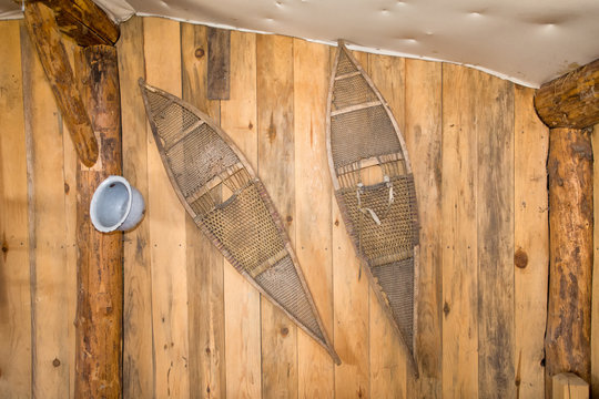 A pair of vintage snowshoes hanging on a wood plank wall supported by raw logs in an old house