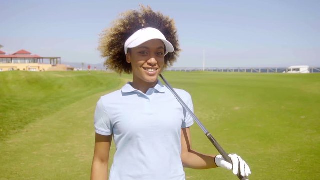 Smiling friendly young African American woman golfer standing smiling happily at the camera with a golf club over her shoulder on a fairway on the course.