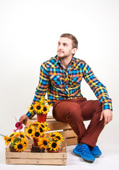 Young florist on a white background. Handsome young man in bright clothes sits on a wooden box with sunflowers in their hands. Care for flowers