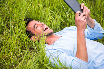 Young happy business man smiling and lying on the green grass looking on device