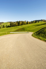 Kurvene Asphaltstrasse durch eine Hügellandschaft im Frühling mit saftigen grünen Wiesen im Entlebuch (Biosphäre) in der Schweiz
