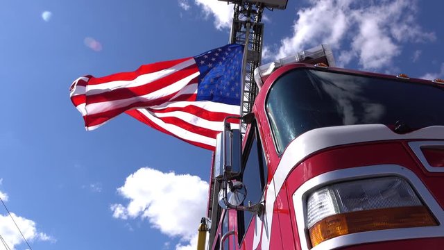 Parked red emergency response vehicle holds waving American flag from ladder under blue sky.
