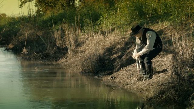 A cowboy from the American wild west era standing next to a creek with a canteen fires his pistol at something in the water. 