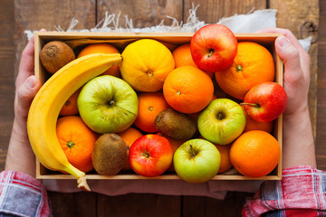 Man in tartan plaid shirt holds a box full of fresh fruits. Fruit harvest - apples, oranges, lemon, banana, kiwi.