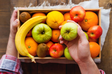 Man in tartan plaid shirt holds a box full of fresh fruits and an apple.