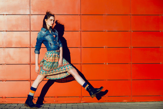 Young teenager girl posing, going, runs near urban red wall background in skirt and jeans jacket on the sunset.